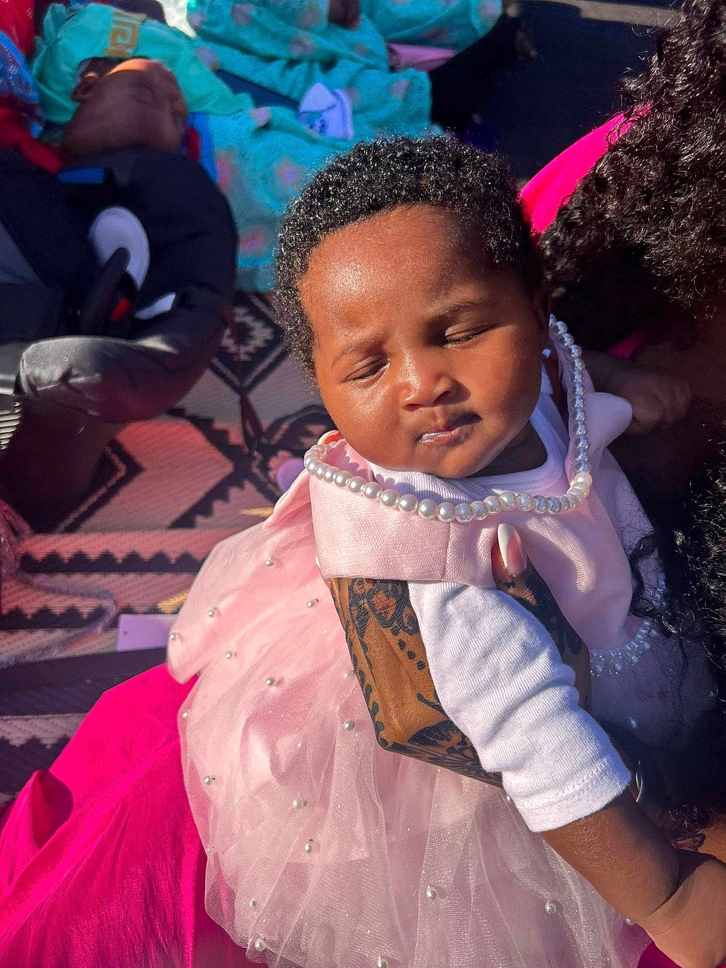 Mariama B is registered to the contest to win money with this photo: baby, child, pink_dress, pearls, tattoo, sleeping, blanket, sunlight, outdoor, person, hand, curly_hair, patterned_rug, clothing, peaceful, infant, resting, portrait, family, holding