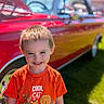 automobile, boy, cartoon_print, casual_clothing, child, cute, daylight, grass, happy, kid, orange_shirt, outdoor, park, portrait, red_car, short_hair, smile, sunny_day, vehicle, vintage_car