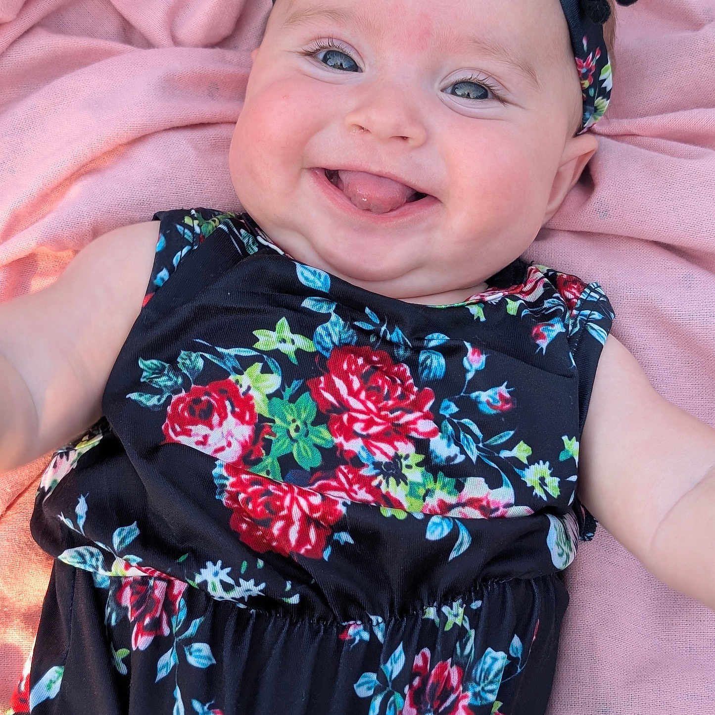 Jaylee is registered to the contest to win money with this photo: baby, smiling, tongue_out, headband, floral_dress, pink_blanket, close_up, child, happy, cheeks, cute, infant, portrait, skin, eyes, person, laying_down, clothing, fashion, soft_texture
