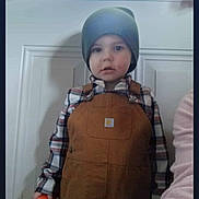 Atrius is registered to the contest to win money with this photo: child, beanie, overalls, plaid_shirt, indoor, door, window, holding_object, orange_object, curious_expression, young_boy, casual_clothing, portrait, person, standing, light_shadow, home_interior, hat, neutral_expression, clothing