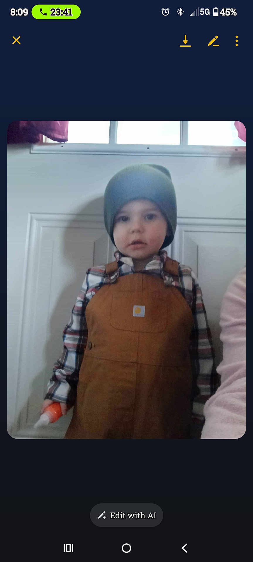 Atrius is registered to the contest to win money with this photo: child, beanie, overalls, plaid_shirt, indoor, door, window, holding_object, orange_object, curious_expression, young_boy, casual_clothing, portrait, person, standing, light_shadow, home_interior, hat, neutral_expression, clothing