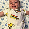 baby, infant, smiling, tongue_out, onesie, truck_print, blanket, teddy_bear_pattern, lying_down, happy, cute, child, playful, indoors, carpet, light_skin, short_hair, portrait, young_child, expressive_face