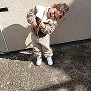 Nolan a rejoint le concours — aidez-le/la à gagner de superbes lots ! child, toddler, smiling, laughing, fleece_jacket, fleece_pants, white_shoes, outdoor, sunlight, shadow, concrete, wall, curly_hair, standing, happy, casual_clothing, daylight, playful, joy, cute