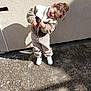child, toddler, smiling, laughing, fleece_jacket, fleece_pants, white_shoes, outdoor, sunlight, shadow, concrete, wall, curly_hair, standing, happy, casual_clothing, daylight, playful, joy, cute