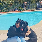 Mia participe au concours pour gagner de l'argent avec cette photo : woman, person, dog, black_dog, large_dog, pet, companion, cuddling, sitting, outdoor, swimming_pool, poolside, garden, backyard, sunlight, summer, casual_clothing, sneakers, tongue_out, portrait