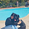 woman, person, dog, black_dog, large_dog, pet, companion, cuddling, sitting, outdoor, swimming_pool, poolside, garden, backyard, sunlight, summer, casual_clothing, sneakers, tongue_out, portrait