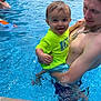 child, toddler, man, swimming_pool, water, outdoor, summer, smiling, tongue_out, playful, holding, family, fun, casual, bright_clothing, necklace, water_splash, backyard, trees, house