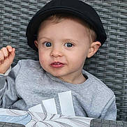 Thibault a rejoint le concours — aidez-le/la à gagner de superbes lots ! child, toddler, cap, hat, blue_eyes, face, sweatshirt, grey_clothing, tablecloth, pattern, hand, expression, portrait, indoor, cute, person, young_child, closeup, seat, woven_background