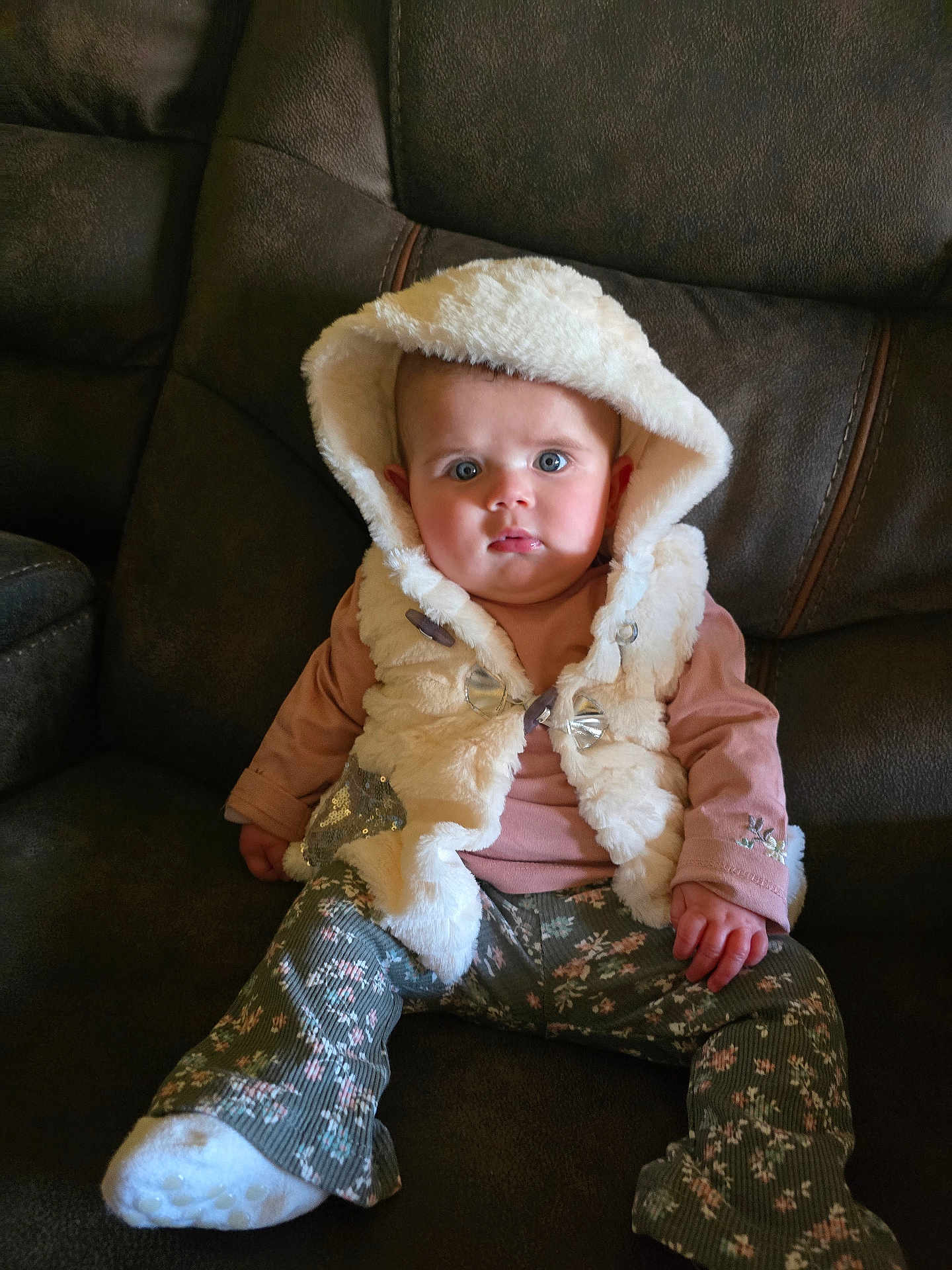 Emberly is registered to the contest to win money with this photo: baby, child, hood, vest, floral_pants, sitting, couch, leather, indoor, cute, face, blue_eyes, clothing, sock, pink_shirt, portrait, young_child, furniture, expression, cozy