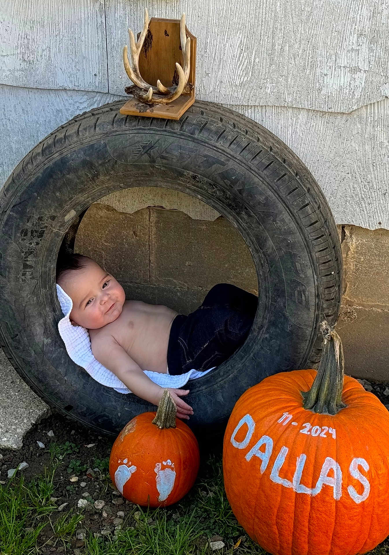Dallas is registered to the contest to win money with this photo: baby, tire, pumpkin, painted_pumpkin, footprints, outdoor, grass, wall, antlers, child, shirtless, jeans, smile, face, head, nature, decoration, fall, autumn, cute