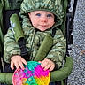 toddler, child, blue_eyes, camouflage_jacket, hood, stroller, toy, pop_it, colorful, outdoor, gravel, hands, cute, infant, jacket, seatbelt, playful, young_child, casual, winter_wear