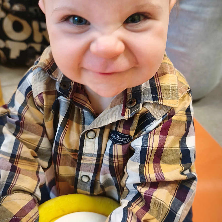 Guerlin a rejoint le concours — aidez-le/la à gagner de superbes lots ! toddler, child, plaid_shirt, toy, drum, hands, face, blue_eyes, candid, indoor, expression, playful, scrunched_face, short_hair, casual_clothing, person, sitting, colorful, orange_surface, cute