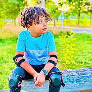 Aayan a rejoint le concours — aidez-le/la à gagner de superbes lots ! child, curly_hair, blue_shirt, knee_pads, elbow_pads, watch, bench, outdoor, greenery, trees, park, sitting, casual_clothing, sandals, pavement, daylight, nature, protective_gear, young, relaxed