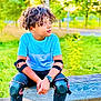 child, curly_hair, blue_shirt, knee_pads, elbow_pads, watch, bench, outdoor, greenery, trees, park, sitting, casual_clothing, sandals, pavement, daylight, nature, protective_gear, young, relaxed