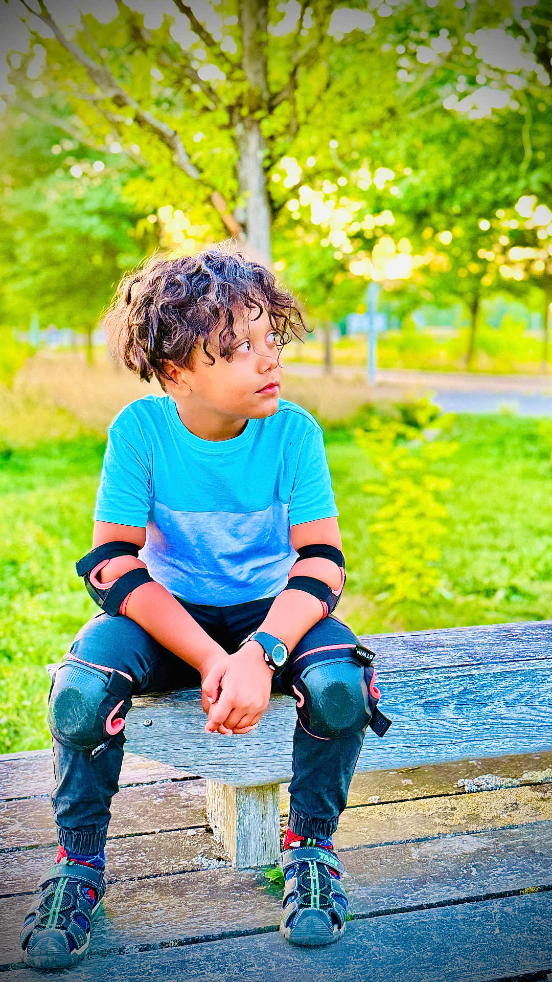 Aayan a rejoint le concours — aidez-le/la à gagner de superbes lots ! child, curly_hair, blue_shirt, knee_pads, elbow_pads, watch, bench, outdoor, greenery, trees, park, sitting, casual_clothing, sandals, pavement, daylight, nature, protective_gear, young, relaxed