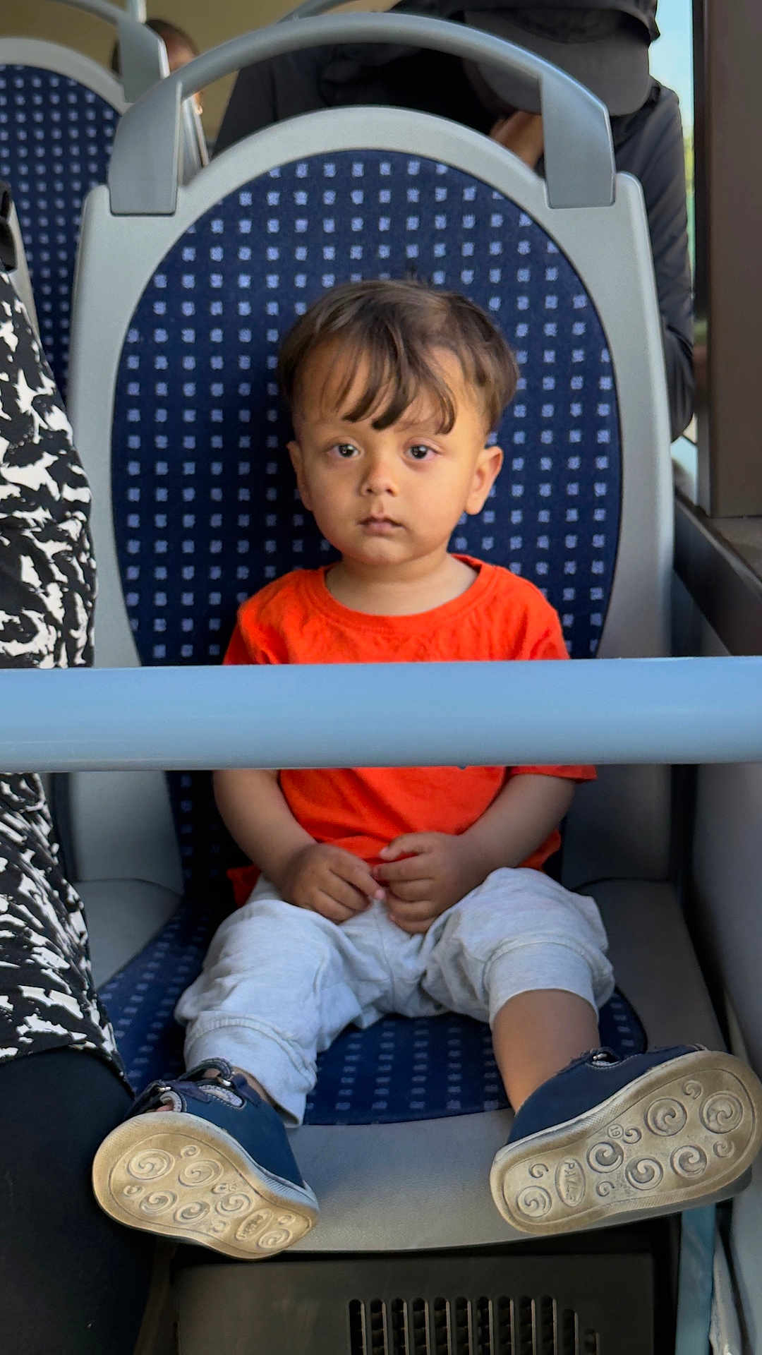 Azlan joined the competition — help win amazing prizes! toddler, child, orange_shirt, white_pants, blue_shoes, seat, public_transport, bus, serious_expression, sitting, safety_bar, sole_of_shoes, patterned_seat, indoor, person, young_child, casual_clothing, short_hair, daylight, travel