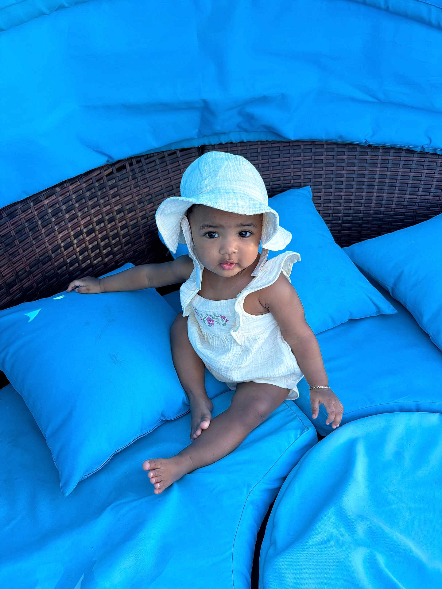 Mylani joined the competition — help win amazing prizes! baby, child, white_hat, romper, blue_cushions, outdoor, wicker_furniture, sitting, curious, infant, sunhat, barefoot, bracelet, plush, comfort, portrait, cute, young_child, relaxing, daylight