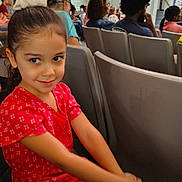 Navia is registered to the contest to win money with this photo: child, girl, red_dress, scrunchie, smile, auditorium, chair, people, seated, crowd, indoor, event, spectators, curly_hair, casual_clothing, portrait, happy, young, cute, person