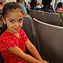 child, girl, red_dress, scrunchie, smile, auditorium, chair, people, seated, crowd, indoor, event, spectators, curly_hair, casual_clothing, portrait, happy, young, cute, person