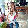 child, girl, curly_hair, tank_top, american_flag, jeans, sandals, watermelon, porch, wooden_floor, table, plants, person, outdoor, smile, pose, hair_bows, rug, casual_clothing, daylight