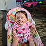 baby, child, smiling, pink_clothing, strawberry_pattern, baby_chair, floral_pattern, indoor, flooring, toy_car, high_chair, happy, cute, person, seatbelt, wood_floor, playful, home, infant, furniture