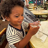 Maylone participe au concours pour gagner de l'argent avec cette photo : child, toddler, curly_hair, striped_shirt, high_chair, glass, empty_glass, table, outdoor_cafe, smile, person, wooden_chair, napkin, daylight, restaurant, background_people, happy, young_child, casual_clothing, hands