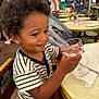 child, toddler, curly_hair, striped_shirt, high_chair, glass, empty_glass, table, outdoor_cafe, smile, person, wooden_chair, napkin, daylight, restaurant, background_people, happy, young_child, casual_clothing, hands
