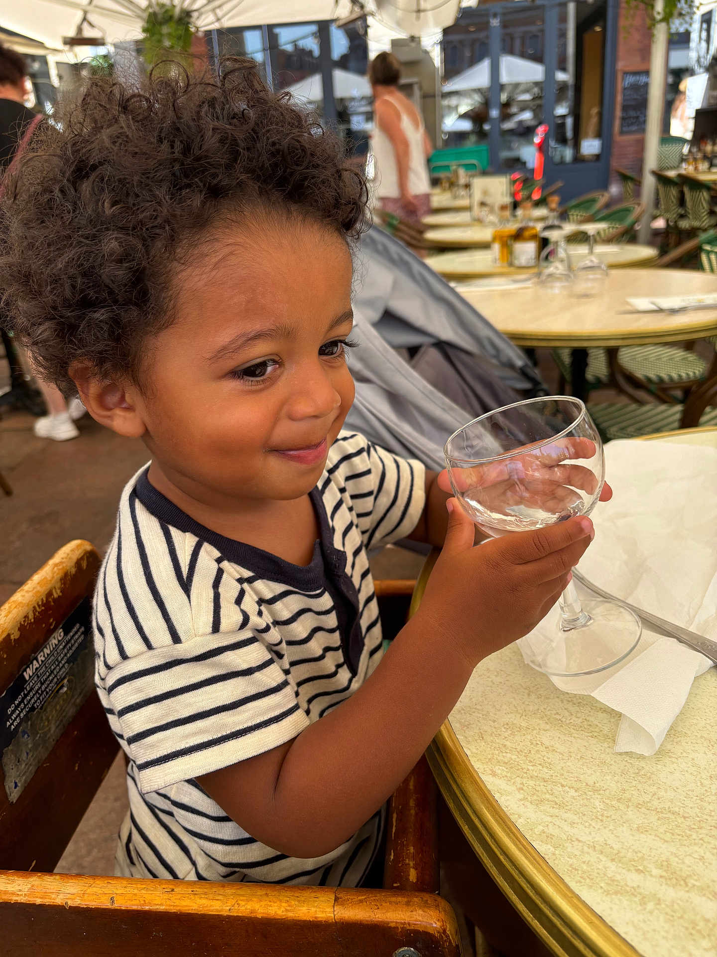 Maylone participe au concours pour gagner de l'argent avec cette photo : child, toddler, curly_hair, striped_shirt, high_chair, glass, empty_glass, table, outdoor_cafe, smile, person, wooden_chair, napkin, daylight, restaurant, background_people, happy, young_child, casual_clothing, hands