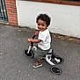 Maylone participe au concours pour gagner de l'argent avec cette photo : toddler, child, smiling, balance_bike, curly_hair, white_shirt, green_shorts, sandals, outdoor, pavement, wall, brick, fence, greenery, person, happy, play, street, vehicle, footwear