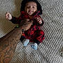 baby, infant, child, carpet, tattoo, hand, socks, hoodie, red_clothing, black_clothing, person, cute, smile, tongue_out, indoor, floor, texture, human, young_child, holding