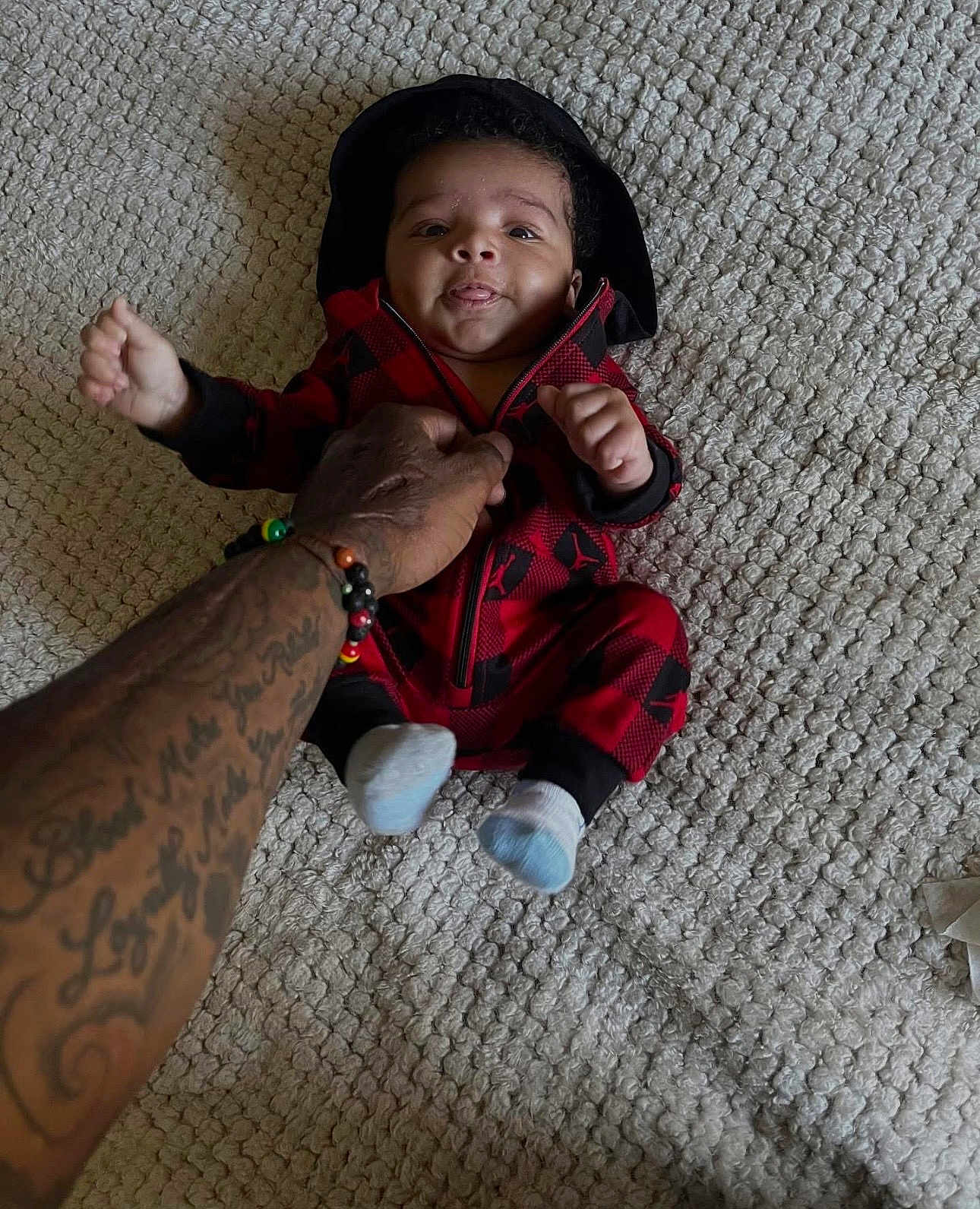 Kealo joined the competition — help win amazing prizes! baby, infant, child, carpet, tattoo, hand, socks, hoodie, red_clothing, black_clothing, person, cute, smile, tongue_out, indoor, floor, texture, human, young_child, holding