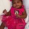 baby, infant, child, portrait, sitting, lying_down, smiling, pink_outfit, dress, skirt, hair_bow, headband, handbag, minnie_mouse, sparkles, socks, bed, bedding, indoors, fashion