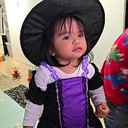 Amarah a rejoint le concours — aidez-le/la à gagner de superbes lots ! child, indoor, costume, witch_hat, purple_dress, black_dress, toy, floor, carpet, curious, standing, person, home, cute, smile, long_sleeve, clothing, hair, face, portrait