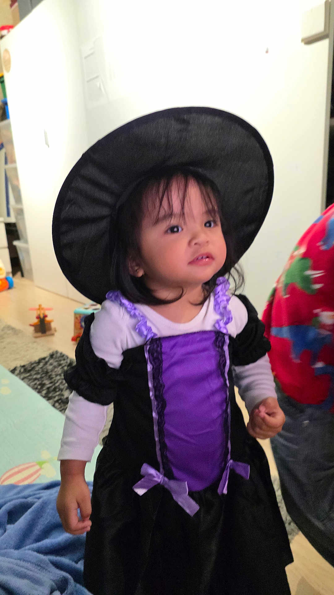 Amarah a rejoint le concours — aidez-le/la à gagner de superbes lots ! child, indoor, costume, witch_hat, purple_dress, black_dress, toy, floor, carpet, curious, standing, person, home, cute, smile, long_sleeve, clothing, hair, face, portrait