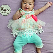 Aria joined the competition — help win amazing prizes! baby, infant, smiling, lying_down, purple_blanket, floral_dress, lace, turquoise_pants, barefoot, milestone_marker, wooden_plaque, happy, cute, child, person, portrait, indoors, soft_texture, celebration, three_months