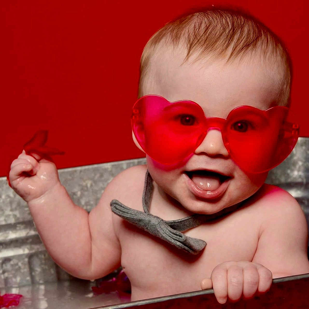 Alex Jr. is registered to the contest to win money with this photo: baby, bow_tie, child, cute, face, fun, hand, happy, heart_shaped_glasses, infant, metal_tub, playful, portrait, red_background, red_glasses, smiling, splashing, summer, tongue_out, water