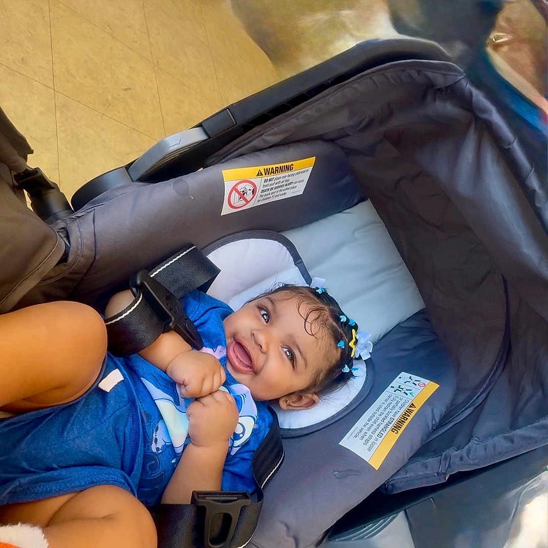 Khari is registered to the contest to win money with this photo: baby, child, infant, smiling, happy, portrait, car_seat, stroller, blue_outfit, hair_clips, curly_hair, sitting, indoors, travel, safety_harness, seat_belt, baby_gear, close_up, photography, family