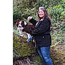 woman, dog, leash, forest, tree, moss, log, outdoor, nature, greenery, smile, casual_clothing, jeans, jacket, pet, animal, happy, standing, canine, walking