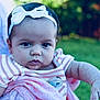 baby, child, headband, bow, pink_clothing, striped_clothing, outdoor, green_background, portrait, infant, cute, face, person, skin, arm, hand, expression, soft_light, natural_light, holding