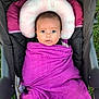 baby, infant, swaddled, purple_blanket, stroller, headrest, pink, crown_pattern, outdoor, grass, child, person, face, eyes, curious, wrapped, seated, comfort, closeup, portrait