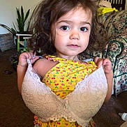 Aubrie is registered to the contest to win money with this photo: toddler, child, bra, yellow_outfit, curly_hair, indoor, living_room, carpet, sofa, plant, furniture, portrait, baby_bottle, expression, clothing, cute, person, face, playful, home