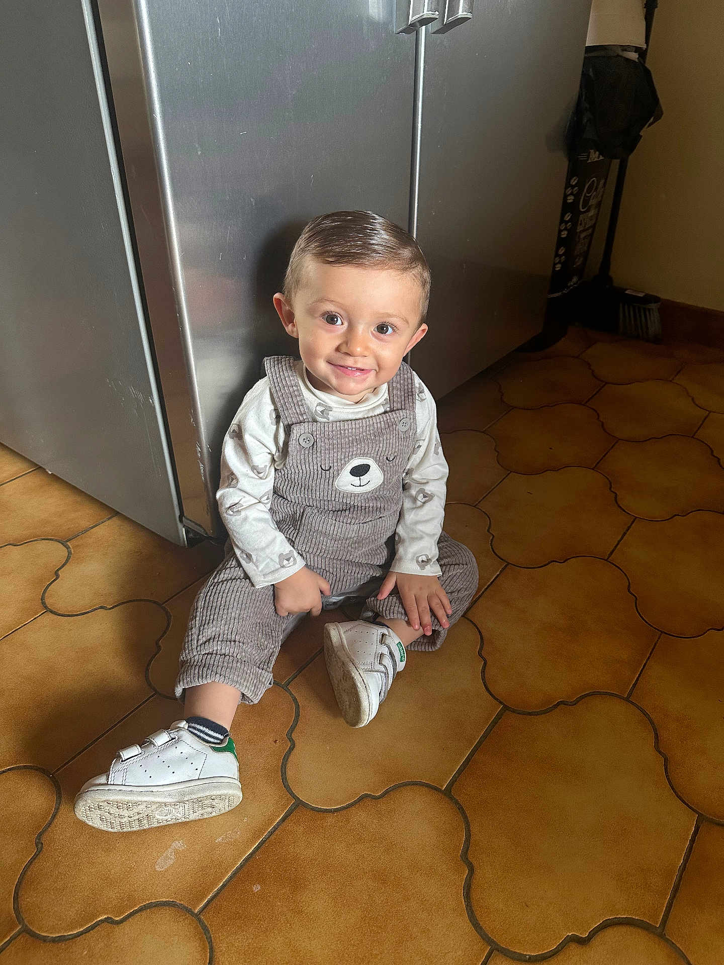 Lélyo a rejoint le concours — aidez-le/la à gagner de superbes lots ! toddler, child, smile, overalls, bear_face, white_sneakers, sitting, floor_tiles, kitchen, patterned_floor, hair, cute, happy, indoor, floor, clothing, person, portrait, young_child, baby
