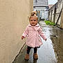 toddler, child, pink_coat, standing, wet_ground, alleyway, puddle, outdoor, rainy, boots, topknot, smiling, concrete, building, wall, grass, satellite_dish, door, person, daylight