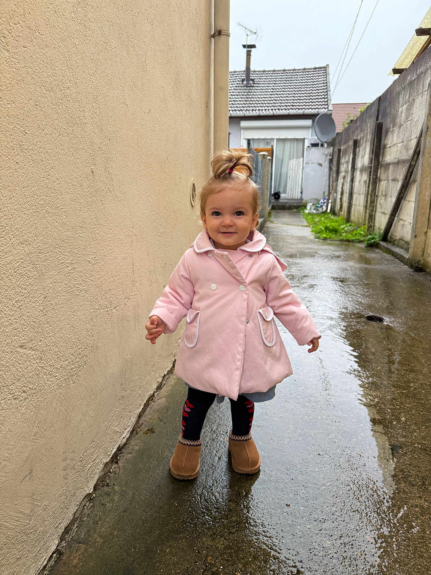 Alice a rejoint le concours — aidez-le/la à gagner de superbes lots ! toddler, child, pink_coat, standing, wet_ground, alleyway, puddle, outdoor, rainy, boots, topknot, smiling, concrete, building, wall, grass, satellite_dish, door, person, daylight