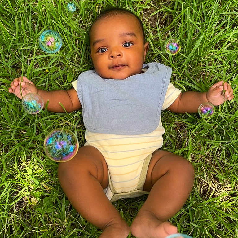 Kenai is registered to the contest to win money with this photo: baby, grass, bubbles, outdoor, infant, blue_bib, onesie, greenery, child, cute, smiling, lying_down, nature, playful, summer, sunlight, happy, peaceful, portrait, closeup
