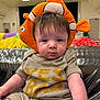 baby, child, plush_hat, clownfish_hat, striped_clothing, indoor, colorful_background, seated, cute, face, headwear, infant, playful, soft_toy, expression, person, short_hair, light, fabric, room