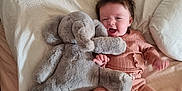 Edie is registered to the contest to win money with this photo: baby, infant, plush_toy, elephant_toy, bed, pillow, blanket, smiling, happy, child, soft_toys, cozy, cute, toddler, clothing, lying_down, indoors, person, sleepwear, furniture