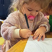 Milla participe au concours pour gagner de l'argent avec cette photo : child, toddler, pacifier, drawing, marker, wooden_table, paper, scribbles, pen, sweater, indoor, hair, messy_hair, concentration, portrait, cabinet, microwave, furniture, tabletop, hands