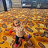 toddler, child, indoor, play_area, colorful, carpet, chocolate_chip_cookie_pattern, balls, pink_socks, strawberry_shirt, happy, smiling, fence, plants, windows, floor, person, young_child, cute, playful