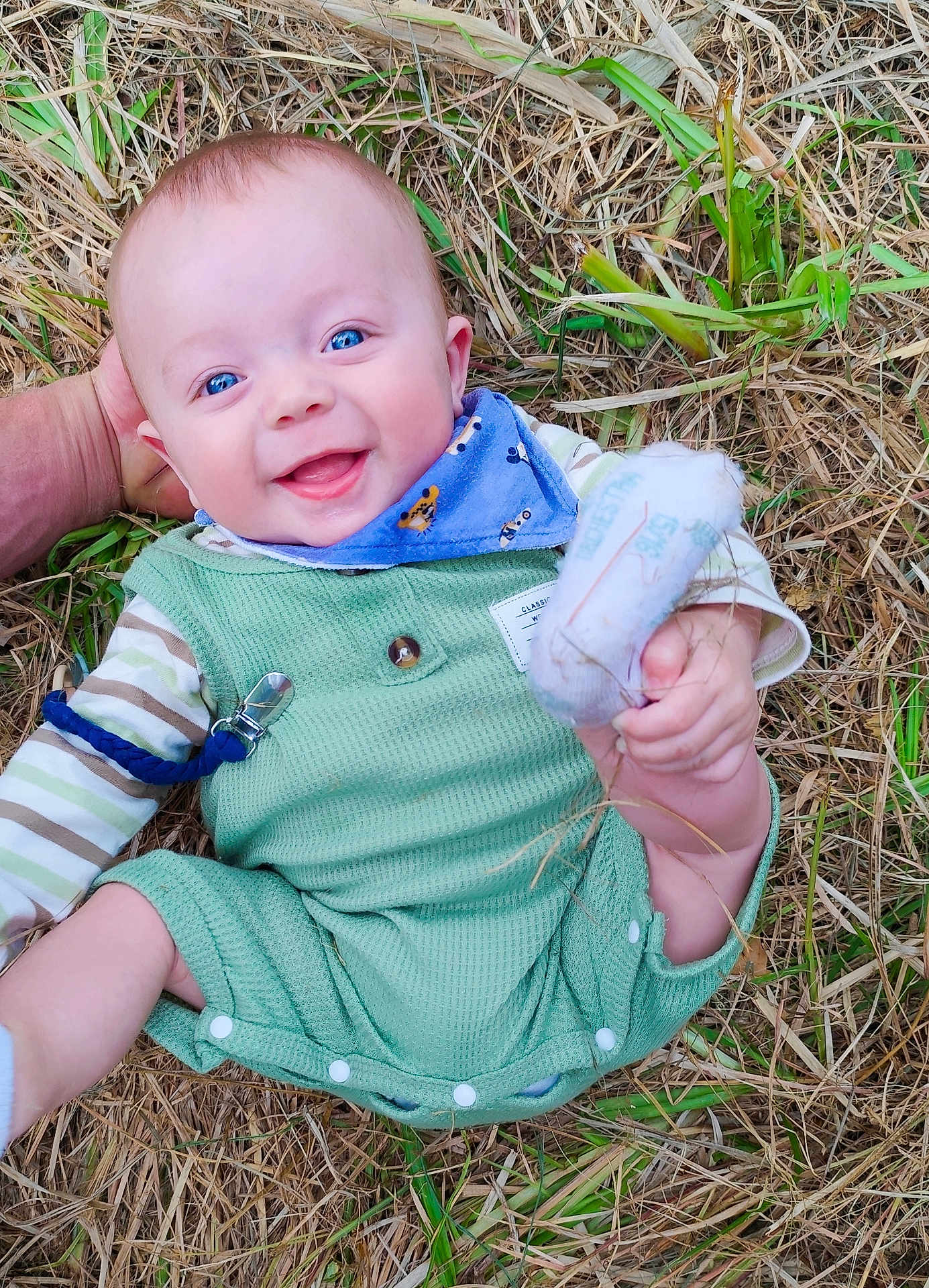 Oban participe au concours pour gagner de l'argent avec cette photo : baby, infant, smiling, blue_eyes, green_clothing, bib, sock, hand, straw, grass, outdoor, cute, happy, person, portrait, lying_down, child, head_support, nature, playful